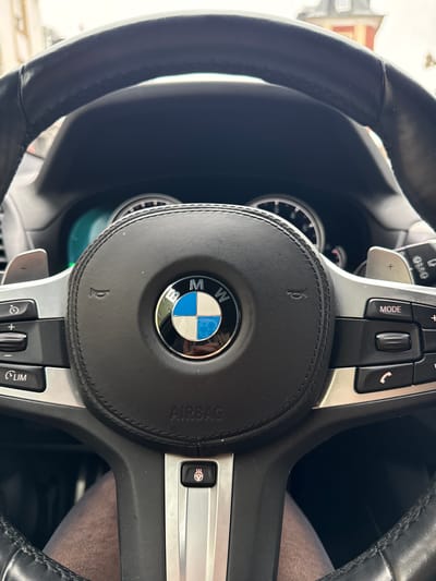 The image presents a driver's perspective from within a BMW vehicle in Pétange, Luxembourg, focusing on the steering wheel and dashboard. The black leather-wrapped steering wheel features a prominent BMW logo at its center, with "AIRBAG" printed below. Silver-colored spokes extend from the hub, equipped with multiple control buttons: the left spoke includes cruise control functions such as speed adjustment and a "LIM" button, while the right spoke contains controls for media (e.g., "MODE") and phone functions. Paddle shifters are visible behind both spokes. A dedicated button with a heating element icon is present on the lower right spoke. The instrument cluster is partially visible behind the steering wheel, displaying a blue-green illumination. The driver's legs, clad in light brown fabric, are visible below the steering wheel. A reflection of a person, likely the driver, wearing a red garment, is discernible within the chrome ring of the BMW emblem. Through the windshield, an out-of-focus street scene of Pétange, Luxembourg, is visible, showing residential buildings with multi-story facades and a red-tiled roof under an overcast sky.