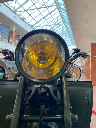 The image is a close-up, eye-level shot of the headlight of a vintage motorcycle, likely on display indoors. The motorcycle is painted a dark green, and the headlight is round with a yellow-tinted lens. The background shows the interior of a building with a glass ceiling, suggesting a museum or exhibition hall. The architecture in the background, with its tall pillars and balconies, is consistent with the Museum of Military Technology in Verkhnyaya Pyshma, Russia. There are no people visible in the frame.