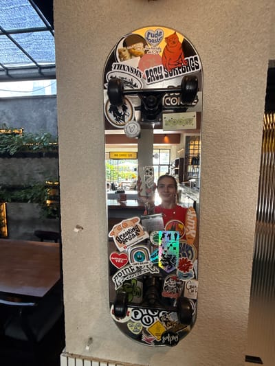 The image shows a reflective skateboard mounted on a textured wall. The skateboard is covered with various stickers. A young woman is reflected in the skateboard's surface, taking a picture of it with her smartphone. The background reflection shows an interior with a window and plants. Based on the stickers and the general aesthetic, the location is Uluwatu, Indonesia.