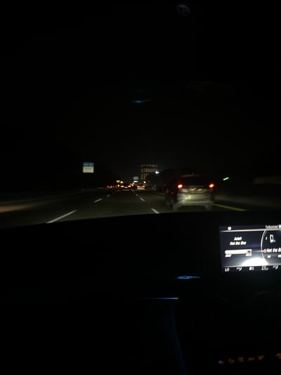 The image shows a nighttime view from inside a car driving on a highway. The road is visible with lane markings and other cars in front, their headlights and taillights illuminated. A digital display screen in the lower right corner of the frame shows song information, with "Asiah" and "Not the One" visible. The background is dark, with some distant lights and buildings that suggest an urban or suburban environment. The overall scene depicts a typical highway journey at night. Without specific landmarks or context, it is not possible to definitively identify the location as Babakan Madang, Indonesia.