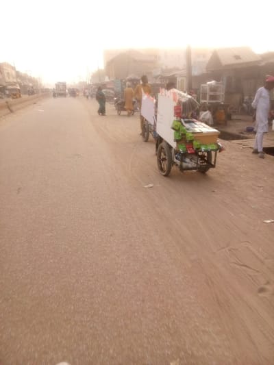 A dusty street scene features a mobile vendor cart laden with snacks. The cart, painted white, has two large wheels and is being pushed along a paved road. Other pedestrians and vendors are visible in the background, adding to the bustling street atmosphere. Buildings line the road, creating a sense of urban density and activity. The lighting suggests a hazy or overcast day, with a yellowish hue permeating the scene.