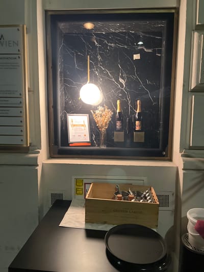 The image shows a dimly lit interior space with a black marble wall, a wooden box with the brand "GRAUD LAROSE" on it, and a large, empty, round, black tray on a black table. The wooden box contains several metal containers, likely for condiments or utensils. Two bottles of wine, possibly champagne, are displayed in the background, illuminated by a hanging light fixture, positioned in front of a black marble wall. The lighting is soft and warm, casting shadows on the objects. The scene likely depicts a restaurant or bar, with the displayed bottles and the condiments suggesting it is a place where alcohol and food are served. The location is a building located at coordinates 48.201, 16.362 in Wien, Austria, suggesting it is located in a busy part of the city.