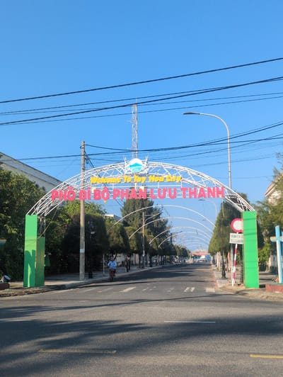 An eye-level outdoor shot depicts an archway over a road, with text in Vietnamese, "Welcome To Tuy Hòa City ĐẢO BẠCH TUYẾT THANH". The archway is constructed of white lattice with red and yellow lettering, with a blue and white emblem.
The scene is located on a street in Tuy Hòa, Vietnam, with the perspective looking down the road. The road is asphalt, with a yellow line on the right and painted white lane markers.
Power lines are visible in the upper left against a clear blue sky. Street lamps stand to the side. Green rectangular structures hold up the arch. Trees and foliage are visible on either side of the road beyond the arch. A motorcyclist is on the left side of the road.