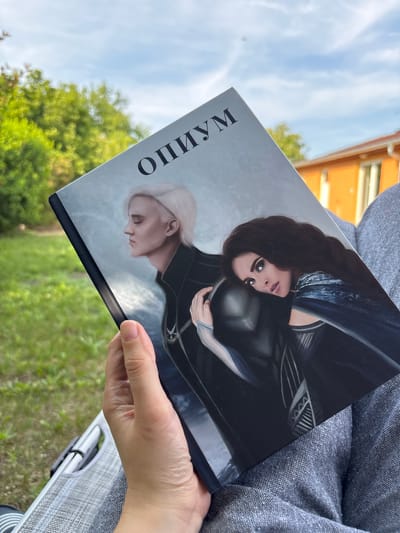A vertical medium shot shows a person holding a book with a cover featuring two illustrated characters. The character on the left has long dark hair and is looking towards the viewer with large, expressive eyes. She is wearing dark armor with blue accents. The character on the right has short, light-colored hair and is looking down with a profile view. This character is also wearing dark attire. The cover is mostly dark, with a gray background behind the characters. To the left of the characters, in large black serif font, is the word "МАЛИПНО". The background of the photograph shows a blurred view of green foliage and a bright blue sky with wispy white clouds. In the upper left corner, out of focus, is a building with an orange roof. The lighting suggests it is daytime, likely sunny. The book is held in the foreground by a person's hand and arm, which are partially visible on the right side of the frame. The person is wearing a textured gray garment. The overall scene appears to be outdoors in a natural setting, possibly a park or garden.