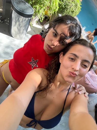 A wide-angle, slightly elevated selfie captures two young adult females posing outdoors next to a swimming pool in Binyamina - Givat Ada, Israel. The female in the foreground, with wet, dark, curly hair, is wearing a dark blue bikini top and is pouting her lips while looking at the camera. Her right arm extends forward to hold the camera, while her left hand rests on the right shoulder of the second female. This second female, positioned behind and slightly to the left, has dark, curly hair and is wearing a red cropped long-sleeved top with a leopard-print star appliqué on the left chest, paired with yellow string bikini bottoms. She looks directly at the camera with a neutral expression.
In the background, a blue swimming pool is visible to the right, showing ripples on the water's surface. A third individual, likely female, in a dark bikini top, is partially visible in the background near the pool edge, next to two clear drinking glasses. To the upper left, a reflective stainless steel cylindrical trash receptacle with a black lid is present on a light-colored paved surface, which displays water reflections. Lush green foliage and potted plants are visible behind the trash can and along the upper right background, indicating a garden or patio setting. Direct sunlight illuminates the scene, creating visible shadows. A light-colored outdoor lounge cushion is partially visible in the lower right corner.