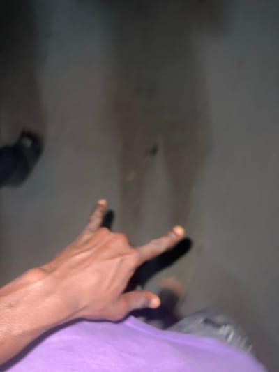 An image captured in Birnin Kebbi, Nigeria, depicts a human left hand in the foreground making the "rock on" gesture (index and pinky fingers extended, middle and ring fingers curled, thumb extended). The hand exhibits dark skin tone and is partially obscured by a light purple garment at the wrist. Directly behind and below the hand, two distinct dark shadows are cast on a light grey-brown, possibly concrete or asphalt, ground surface. These shadows are elongated and appear to be cast by human figures, with one shadow suggesting a torso and leg outline. The environment is dimly lit, and the ground surface shows minor discolored marks. No other subjects or objects are distinctly identifiable.