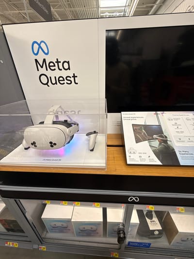 This media file captures a retail display for Meta Quest virtual reality headsets in what appears to be an electronics section of a store in Worcester, United States. The image is oriented vertically, showcasing a well-lit product presentation.

The central focus is a white Meta Quest 3S headset, encased within a clear acrylic display stand. The headset features three black circular sensors on its front and is accompanied by two white Meta Quest controllers, one positioned beside it and the other slightly above. The display base illuminates the headset with soft blue and pink light. A large white panel to the left prominently features the blue Meta logo (an infinity symbol) and the words "Meta Quest" in black text.

Above the physical product display, a large, dark display screen is off, reflecting overhead lighting. To its right, a promotional poster or part of the product packaging highlights the Meta Quest 3S, featuring an image of a person wearing the headset. This graphic advertises "Unreal experiences. Unreal price." and "So. Many. Experiences."

Adjacent to the main display, a glass-fronted retail cabinet holds multiple boxed Meta Quest 3S units, indicating available stock for purchase. Yellow price tags are visible on some of these boxes. Also within this cabinet, a black "onn." branded box, possibly an accessory like a carrying case, is visible. The overall setting suggests a modern retail environment, with metal shelving structures visible in the background on the far left.