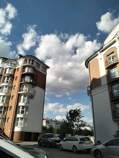 The image depicts a street scene in Krykhivtsi, Ukraine. Two buildings, one on the left and one on the right, frame the view. The building on the left has a white and brown facade, multiple windows, and rounded balconies. The building on the right has a white and brown facade, with rounded corners, windows, and balconies. A blue sky with puffy white clouds is visible between the buildings. In the foreground, several cars are parked along the street. In the background, other buildings are visible amidst trees. The image was taken during the daytime.