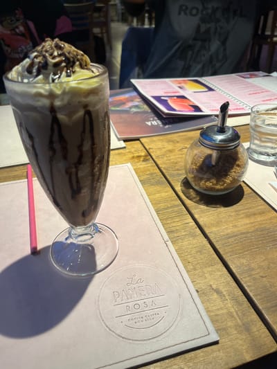 The image depicts a table setting at "La Panera Rosa" restaurant in Lomas de Zamora, Argentina. A chocolate milkshake, topped with whipped cream and chocolate drizzle, sits on a pink placemat. A pink straw rests beside the milkshake glass. A sugar dispenser filled with brown sugar and a glass are positioned on the wooden table, along with a menu. A person wearing a "Rock" t-shirt is partially visible in the background.