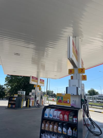 The image shows a Shell gas station with multiple fuel pumps and a canopy. A display rack with various motor oils and antifreeze is in the foreground. The gas station is equipped with a digital display showing fuel prices, and signage indicating different fuel types. There are no people visible in the image, but there is a poster with a person's image on it. The environment appears to be a typical urban setting with trees, roads, and buildings in the background. The sky is clear and blue. This gas station is located in Komotini, Greece.