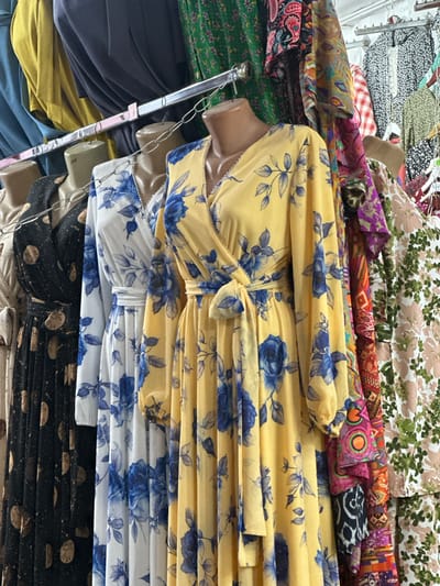 The image depicts a row of dresses on mannequins displayed in an outdoor market setting in Kara-Suu, Kyrgyzstan. The dresses are of various patterns and colors, with a prominent yellow dress featuring blue floral prints in the foreground. The market appears to be a textile or clothing stall, with racks of clothing visible in the background. The lighting suggests it is daytime. The overall scene is that of a typical retail environment in a Central Asian market.