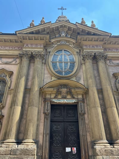 The image shows a close-up, low-angle view of the facade of a church in Turin, Italy. The architecture is ornate, featuring large Corinthian columns, a prominent round window with gridded panes, and carved stone detailing. The building's exterior is a muted pinkish-brown, contrasting with the golden hue of the stone columns and accents.
Above the main entrance, which consists of a large, dark wooden double door with intricate carvings, there is a vertical sign with text that appears to be in Italian. The visible text on the sign reads "INDULGENZA PLENARIA PERPETUA" and partially "OTTA DIANA".
On the left side of the facade, text is carved vertically into the stone, reading "FRANCISCO ASS". On the right side, more text is visible, reading "STIA SACR". Between the columns, there are decorative carvings and what appear to be small statues or gargoyles on the upper corners of the building's pediment. A cross is visible on the far left, mounted on a spire.
The sky is a clear, bright blue, suggesting a sunny day with good weather. The lighting casts distinct shadows, highlighting the architectural depth and texture. The image is a detailed capture of the religious building's exterior, emphasizing its classical design elements.
