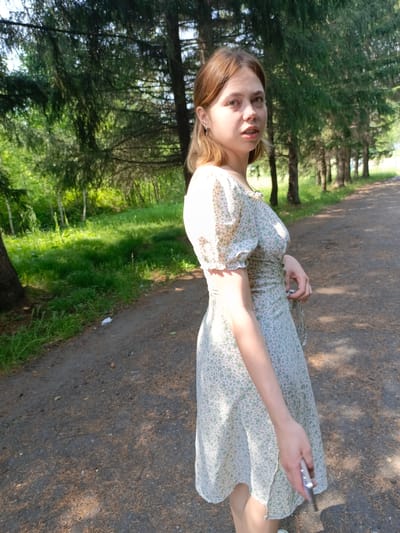 In Zalesovo, Russia, a young woman with light brown hair stands on a dirt path lined with evergreen trees. She is wearing a light-colored floral dress and holding a silver object in her right hand. Sunlight filters through the trees, casting dappled shadows on the path and the surrounding green grass.
