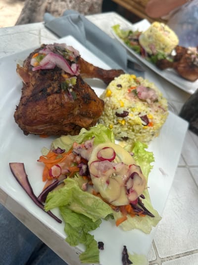 The image shows a plate of food, possibly taken at a restaurant in Le Gosier, France, given the setting. The plate features a piece of grilled or roasted chicken with a garnish of pickled onions and other vegetables. A portion of rice with corn and kidney beans is placed next to the chicken. A salad with lettuce, shredded carrots, sliced radishes, red cabbage, and a creamy dressing is also present on the plate. Another plate of food is partially visible in the background, suggesting that it is a shared meal setting. The table has a textured, possibly tiled, surface.
