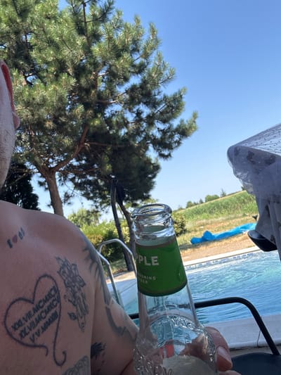 This is a close-up, outdoor, eye-level media file taken on a sunny day, likely in the late morning or early afternoon, given the bright sunlight and clear blue sky. The scene appears to be a poolside or waterfront location in Balchik, Bulgaria, with a swimming pool visible in the background and lush green trees, including a pine tree, framing the shot.
The primary subject is a person's shoulder and arm, covered in several tattoos. One prominent tattoo is a heart shape with Roman numerals inside. Another tattoo depicts a floral design. The person is holding a glass bottle, with its top and neck clearly visible. The bottle has a green label with the word "APPLE" in large white letters and smaller text below that reads "REFRESHING CIDER".
The person seems to be relaxing, possibly enjoying a drink by the water. The overall mood is one of leisure and warm weather enjoyment. In the background, beyond the pool, there's a hint of manicured greenery, suggesting a resort or private residence. A white, patterned fabric, possibly a tablecloth or shade, is also visible at the top of the frame.