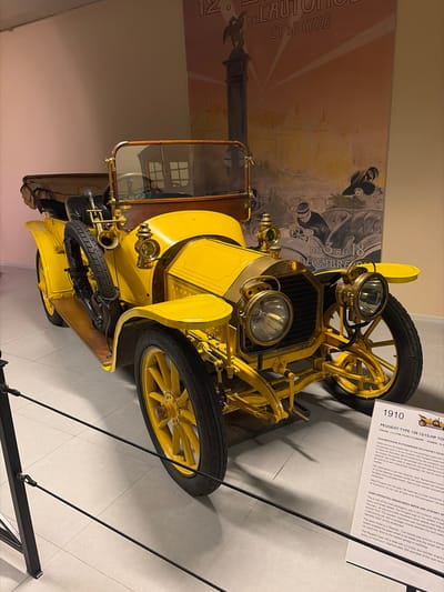 A bright yellow 1910 Peugeot Type 126 12/15-HP touring car is prominently displayed indoors, likely within a museum or exhibition hall in Wassenaar, Netherlands. The vintage vehicle features a vibrant yellow body, large brass headlamps, a brass horn, and wooden spoked wheels, with a spare tire mounted on its side. A wooden-rimmed steering wheel is visible through the windshield.
Behind the car, a large, vintage-style poster is affixed to the wall, partially visible. It reads "12. DE L'AUTO ET DU CYCLE" and "DU 3 AU 18 DÉCEMBRE", depicting stylized figures in period attire, suggesting an early automobile and cycle exhibition.
To the right of the car, a white display board provides detailed information about the Peugeot Type 126. The text, in both Dutch and English, identifies the car as a 1910 model with a 2.2-litre four-cylinder engine producing 15 horsepower. It highlights features like its hand-operated windscreen wiper and Stepney spare wheel system, and mentions that approximately 350 examples of this type were manufactured. The board also notes its aesthetic appeal: "This is one of the most photogenic cars in the museum, partly due to its bright yellow colour and brass details."
The car is positioned on a light-colored tiled floor, with black metal barriers in the foreground indicating a viewing area. The setting is well-lit by artificial lighting, creating a clear and informative display of the historic automobile.