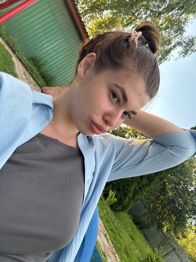 A young woman with brown hair pulled up in a bun is outdoors, with a blue shirt over a grey tank top. She is looking towards the camera, her right arm resting behind her head. The background consists of greenery, a fence, and a portion of a green roof. The lighting suggests it is daytime. The image does not contain information that places it in Мещёры, Russia.