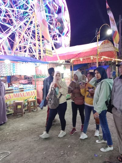The image shows a group of seven people standing in front of a food stall. The stall is located next to a large Ferris wheel with a sign that reads "Di Gunakan Untuk Keluarga" (Used for family). The people are wearing casual clothing. The women are wearing hijabs. Two of the women are holding drinks, while the others are holding phones or bags. The man on the right side of the image is wearing a white shirt and black pants. The ground is covered in dirt. The image is likely taken in a night market in the city of Kaliwungu, Indonesia, based on the provided coordinates. The image is a still shot.