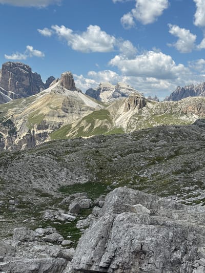 The image depicts a mountainous landscape in the vicinity of Toblach - Dobbiaco, Italy, under a bright, partly cloudy sky. The foreground is dominated by a rocky, uneven terrain with large, grey boulders and patches of sparse green vegetation.

In the mid-ground, the landscape transitions to gentler slopes covered with green grass and interspersed with rocky outcroppings. A group of approximately ten people, appearing as small silhouettes, are visible standing on one of these grassy ridges, looking out towards the mountains.

The background features a series of imposing mountain peaks and ridges with sharp, rugged formations. These mountains exhibit varying textures and colors, ranging from pale grey rock to patches of scree and lighter colored stone. The geology suggests a dramatic, eroded landscape.

The sky is a vibrant blue with scattered cumulus clouds, some of which are illuminated by the sun. The overall impression is of a high-altitude, natural environment. There is a faint inscription, "MESSI", visible on the rocky ground in the mid-ground, to the right of center.