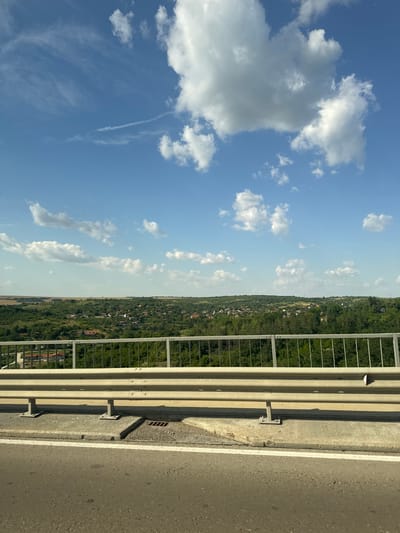 The image is a photograph taken from a bridge, looking out over a rural landscape. In the foreground, there is a section of the bridge's guardrail, made of metal rails and support posts, and the asphalt road surface with a white line marking. Beyond the guardrail, there is a dense green forest that slopes down into a valley. Scattered within the valley and on the distant hills are houses with red roofs, suggesting a village or small town. The sky above is blue with scattered white cumulus clouds. The overall impression is a peaceful, pastoral scene on a bright, sunny day. This scene appears to be located in or near the village of Pisanets, Bulgaria, judging by the architectural style of the houses and the general topography of the region. There are no people or animals visible in the image.