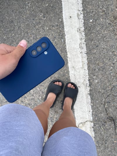 A person is holding a dark blue smartphone in their left hand, positioned over their right thigh which is covered by a light grey pair of shorts. The phone has a triple camera module. The person is wearing black slide sandals on both feet. They are standing on an asphalt surface with a white painted line running vertically along the right side of the frame. The background is a grey asphalt surface with scattered debris and small green blades of grass. The image is taken from a high-angle perspective, looking down at the person's legs and the phone. The lighting suggests it is daytime. The scene appears to be outdoors, possibly on a street or sidewalk in Moskovskoe, Russia.