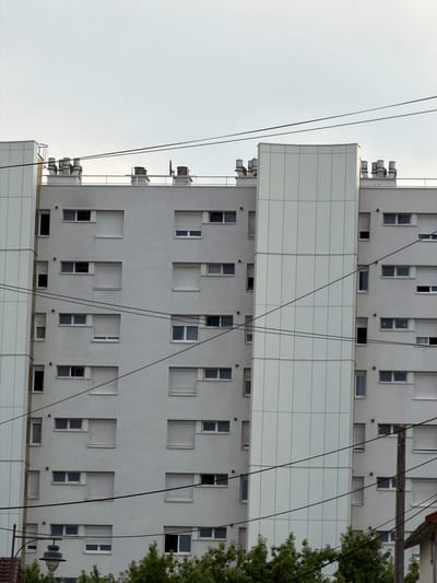 This is a medium shot, taken from a low angle, of a modern, multi-story building. The building is primarily white with a grid-like facade featuring numerous windows of varying sizes. Some windows are dark, suggesting they are closed or unlit, while others appear to be open or reflective. Interspersed with the windows are small, circular ventilation elements.
The building's structure is segmented by horizontal bands of white paneling. Hanging cables, likely power lines or communication wires, are prominently visible in the foreground, crossing diagonally across the frame. On the right side of the image, a lush green tree extends into the frame, its foliage contrasting with the stark white of the building. A street lamp with a dark, round fixture is also visible in the lower right corner, partially obscured by the tree.
The lighting suggests it is daytime, with a bright, overcast sky serving as the background. The overall scene is urban and contemporary. There is no visible text in the image. The absence of people and the static nature of the architecture create a quiet, almost impersonal atmosphere. The orientation of the image is rotated, making the building appear to be on its side.