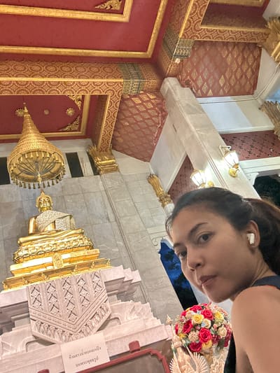 A woman with a ponytail and white earbuds is taking a selfie in front of a large golden Buddha statue. The Buddha is seated on an ornate pedestal, with a decorative umbrella-like canopy above it. The ceiling of the structure is ornate, with red and gold patterns. There is a floral arrangement in front of the pedestal. The background includes stone pillars and lighting fixtures. The scene appears to be inside a temple or religious site in Tha Khlong Town Municipality, Thailand.