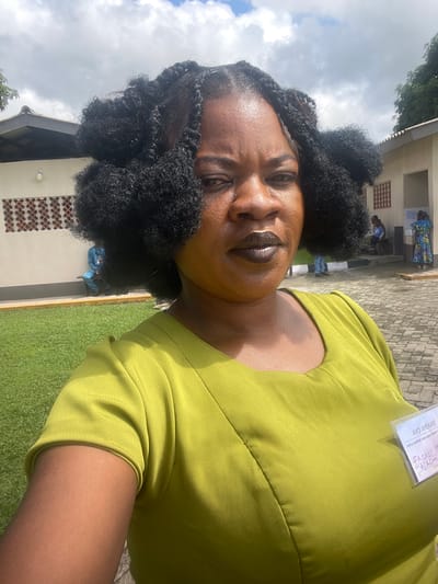 A selfie taken by a Black woman with dark skin and voluminous braided hair. She is wearing a lime green dress and dark lipstick. She has a nametag pinned to her dress. In the background, there is a building with an outdoor area covered in paving stones and a grassy lawn. Several people are visible in the background, some sitting and some standing. One person is wearing a blue uniform, and another is wearing a colorful dress. The sky is cloudy. The image was taken in Itoga, Nigeria.