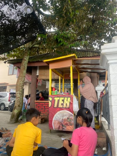 The scene takes place in a food stall in the Tawangmangu district of Indonesia, located at the coordinates -7.669 latitude and 111.111 longitude. The stall is constructed of wood and metal, with a yellow canopy and a sign that reads "Teh" in red letters. There are various items for sale on the counter, including a clear container with what appears to be food. A woman wearing a beige hijab and long dress is standing behind the counter, potentially assisting customers. In the foreground, three individuals are seated on the ground. One is a young boy wearing a yellow shirt, another is a young girl wearing a pink shirt, and the third is partially obscured by the frame. The girl is holding a white object and looking down at it, while the young boy is looking to the side. The area is shaded by a large tree with thick leaves, and the concrete ground is littered with various pieces of debris.