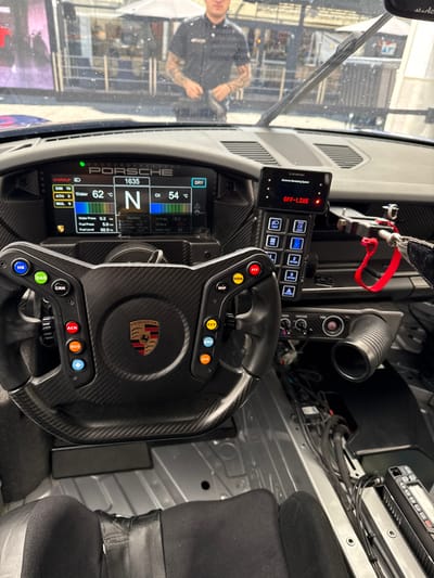 The interior of a Porsche race car with the driver's seat visible. The driver is not in the car, and the steering wheel is centered. The dashboard displays "1635," "62°C" for water temperature, "N," and "54°C" for oil temperature. The car is in a "dry" condition, with the "off-line" message illuminated on the control panel. The dashboard includes many buttons and controls, including "HB," "RAD," "CRK," "ACK," "RVS," "WIP," "MRK," "PIT," "FCY," "D18," and "D16." The car is located in Stavelot, Belgium, at the coordinates 50.441° N, 5.969° E. The environment is not visible. The subject of the image is the interior of the Porsche race car.