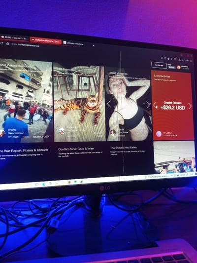 The image displays an LG computer monitor situated on a desk in an indoor environment in Tiruchirappalli, India, illuminated by blue and purple ambient lighting. The monitor screen exhibits several distinct vertical white lines, indicative of potential screen damage or glare. The screen displays the website `collectivememory.ai`, featuring a grid-based interface.

The primary content on the monitor includes three visible "memory" sections:
1.  **"Most Valued Memories":** Features a wide-angle image of a crowded street scene with numerous individuals, some wearing red attire, potentially a protest or gathering. A person in a blue shirt is visible from the back in the foreground. Text below reads "HASAN Gaza, Unknown 7/11/2025" and "$3,058.3 USD," with the section title "The War Report: Russia & Ukraine."
2.  **"Hot Memories":** Presents an image of a tan and black Bengal cat lying on a light-colored wooden floor. Accompanying text indicates "Los Angeles, USA" and "$2,374.0 USD," under the title "Conflict Zone: Gaza & Israel."
3.  **"Latest Memories":** Shows a light-skinned woman with extensive tattoos on her arms and torso, wearing a black sports bra, with one arm raised to her head. Text below identifies her as "Senya Perm, Russia 9/2/2026," and the section title is "The State of the States."

A red sidebar on the right of the screen displays "Latest Activities" and a "Creator Reward +$26.2 USD," along with a timestamp "2/2/2026, 08:48 PM." The Windows taskbar is visible at the bottom of the monitor, displaying various application icons and a partially visible Indonesian text pop-up. In the physical foreground, a cluster of black cables is visible resting on the desk surface in front of the monitor's stand, and the right portion of a white keyboard is also discernible.
