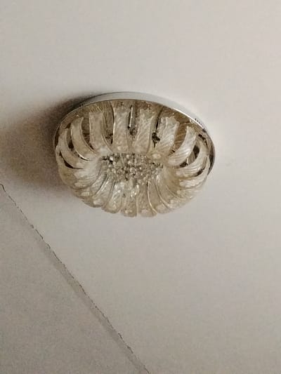 A low-angle, close-up shot depicts a clear glass flower-shaped ceiling light fixture mounted on a white ceiling. The fixture has a chrome or silver-colored trim. A diagonal crack runs across the ceiling on the left side of the image. The location is impossible to determine from the image.