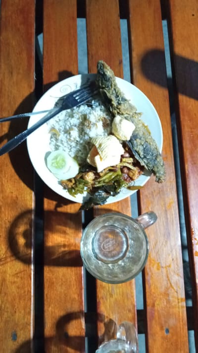 An overhead shot captures a meal on a wooden table in Julicotmeurak, Indonesia. A white plate holds a portion of rice, topped with a fried fish and several crispy crackers. A slice of cucumber and some green vegetables accompany the main dish. A fork rests on the plate. To the right of the plate, a glass of liquid is partially visible. The wooden planks of the table are illuminated by what appears to be artificial lighting, casting shadows on the surface.