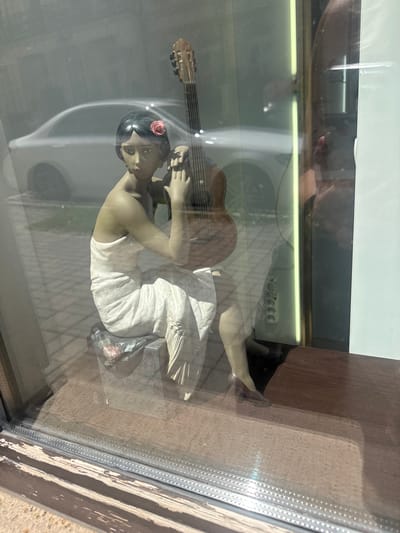 The image shows a figurine of a woman in a white dress playing a guitar. She has dark hair with a red flower behind her ear. The figurine is positioned on a wooden surface, possibly a shelf or display. The background appears to be a window with reflections of a person, a white ceiling, and possibly some lights. The scene is dimly lit, suggesting it could be indoors or twilight. The overall impression is a still life arrangement. The location context is Karlovy Vary, Czechia. There is no visible text in the image.