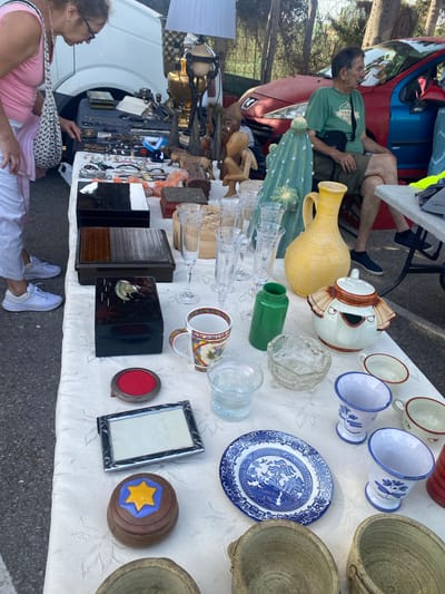 This image depicts an outdoor market stall in San Luis de Sabinillas, Spain, with a variety of items for sale. A woman wearing a pink tank top and white pants is examining items on a table covered with a white cloth. The table displays an assortment of objects including wooden boxes, jewelry, glassware, a ceramic pitcher, a teapot shaped like a face, decorative plates, and various ceramic cups. In the background, a red car is parked, and trees are visible. Another person, a man wearing a green t-shirt and shorts, is seated on a chair near the car, observing the market. The setting suggests a typical flea market or street market scene.