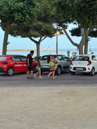 In Sant Feliu de Guíxols, Spain, at sunset, a man wearing a black t-shirt and shorts walks beside a young girl in a light blue skirt and dark t-shirt, who is walking away from the camera. They are near a red car with "GINESTA GA" visible on its side. A woman with long dark hair, wearing a pink dress and carrying a green backpack, walks in the opposite direction of the man and girl, with a toddler on her hip. She is passing a grey car. Behind the grey car is a white Kia Picanto with license plate "7632 LZF". The scene is a paved area beside a body of water, with trees overhead and a streetlamp visible in the background. The sky is a pale blue, indicating dusk.
