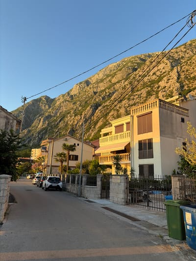 The image shows a street scene in Dobrota, Montenegro, at golden hour. A paved road runs through the center of the frame, lined with stone fences and manicured gardens. Several cars are parked along the side of the road. On the right, two residential buildings are visible, one a pale yellow with balconies and awnings, and another a light beige with dark-shuttered windows. Behind the buildings, a steep, rocky mountain rises dramatically against a clear blue sky. The mountain is partially covered in sparse vegetation. Electrical wires stretch across the upper portion of the image, connecting to a utility pole on the left. A green and a blue trash bin are situated near the sidewalk on the right.