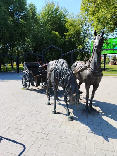 This media file shows a bronze sculpture of a horse-drawn carriage in a park. The sculpture features two horses harnessed to a carriage. One horse is facing forward, while the other is bowing its head. People are seated inside the carriage, and others are standing nearby. The setting is a paved walkway with trees and green grass in the background. A large digital billboard is visible in the distance, displaying text in Russian. The sun is shining, casting shadows on the ground, suggesting it is daytime. The overall atmosphere appears to be relaxed and leisurely, as if people are enjoying a day out in the park. The location context is Dolgoprudny, Russia.