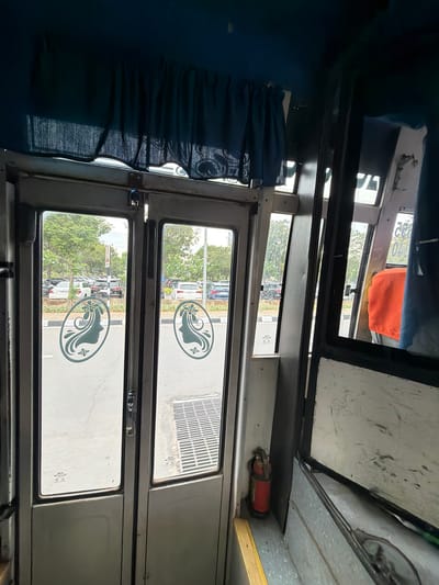 The image shows the interior of a vehicle, likely a bus, looking out through the doors. The doors are glass with a metal frame and feature stylized silhouette decals of women's faces with flowers in their hair. Outside, a parking lot filled with cars is visible, surrounded by trees and a cloudy sky. A fire extinguisher is mounted on the interior wall of the vehicle near the door. The interior of the vehicle appears worn, with some scuff marks and a blue curtain visible at the top. The scene is set in Gollapalle Khurd, India, suggesting a common mode of transport within or around the locality.