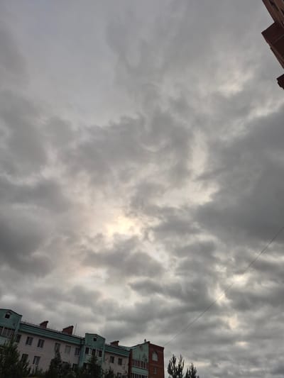 The image captures an overcast sky with dramatic, textured clouds. Sunlight struggles to break through the dense cloud cover, creating a diffused, bright glow in the upper center of the frame. A thin, dark wire stretches diagonally across the sky from the upper right towards the center.
In the lower right and upper left corners, portions of buildings are visible. The building on the right appears to be a multi-story apartment complex with a light-colored facade, green trim around the windows, and darker rooflines. Some windows are visible, along with architectural details like dormer windows. A dark green evergreen tree partially obscures the rightmost part of this building. The building on the upper left is a darker, reddish-brown structure, also appearing to be a multi-story building.
The scene suggests an urban or suburban environment. The time of day is unclear due to the overcast conditions, but the diffuse light implies it is daytime, possibly late afternoon or early evening. The weather is clearly cloudy and appears to be cool or temperate. There is no visible text in the image. The overall mood is somber and perhaps a little dramatic due to the intensity of the clouds. The context is Nyagan, Russia.