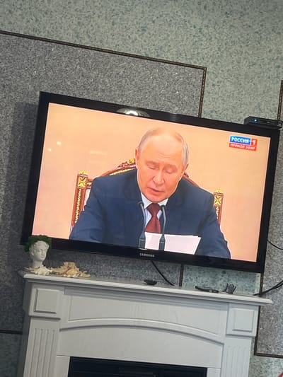 This media file depicts a television screen displaying a news broadcast. The screen shows a close-up of a man, identified by the Russian text "РОССИЯ 1" and "ПРЯМОЙ ЭФИР" (Russia 1, Live Broadcast) in the upper left corner. The man appears to be Vladimir Putin, wearing a dark suit and a red tie, with his eyes closed and head slightly bowed. He is seated at a desk with papers in front of him, and microphones are visible. The background is a plain, light-colored wall, suggesting an indoor setting, likely a studio. The overall mood is serious and formal. The television itself is a Samsung brand, and it is placed on a grey, speckled floor with a dark border. To the right of the television is a white, decorative structure, possibly a fireplace surround, with ornate detailing. There is a small, classical bust with green foliage at its base placed on the edge of the television. The image is captured in what appears to be daytime, given the lighting on the television screen. The setting appears to be a domestic interior in Елецкое, Russia.