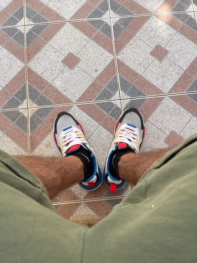 The image shows a pair of feet wearing colorful athletic shoes and black socks, standing on a tiled floor. The tiles are arranged in a diamond pattern with various colors including white, grey, and reddish-brown. The person is wearing olive green shorts. The lighting suggests it is daytime, possibly outdoors or in a well-lit indoor space. The scene is simple and focuses on the footwear and the tiled surface. There is no visible text in the image. The setting appears to be a residential area, given the style of tiling.