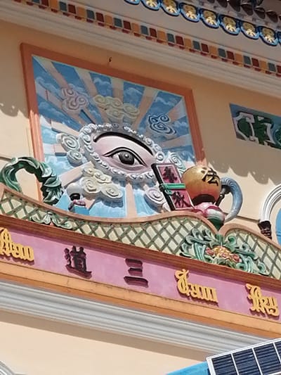 The image shows a detailed close-up of the exterior facade of a building in Lộc Quý, Vietnam. The building's architecture features intricate decorative elements, including a large, stylized eye with swirling patterns surrounding it, rendered in relief against a backdrop of blue and white rays and clouds. To the right of the eye, a golden spherical object with dark Chinese characters is visible, connected to a blue, horn-like protrusion. Below this central motif, the facade displays decorative friezes and inscriptions. A pink band runs horizontally, adorned with yellow, stylized lettering and dark, embossed characters that appear to be in Chinese or Vietnamese. The upper edge of the building has a colorful decorative border with alternating red and yellow square patterns. Green, leafy, sculpted elements are also integrated into the facade's design. The overall impression is one of ornate, possibly religious or culturally significant, architecture.