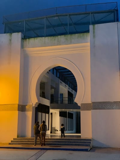 A low-angle shot captures a modern building at dusk. The structure features a prominent archway, revealing an interior courtyard with multiple levels and geometric architectural elements. Two figures stand on the steps leading into the building, while another appears further inside. The building's exterior is illuminated by warm lighting, contrasting with the cool blue tones of the evening sky. A glass-covered structure is visible on the left side. The overall impression is one of contemporary design and understated elegance.
