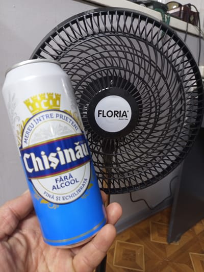 A hand holds a can of "Chisinau" beer in front of a black oscillating fan. The beer can is white with blue and yellow accents and features text in Romanian. The fan has a circular black grille and a white circular logo in the center that reads "FLORIA". The background consists of a gray wall and a brown tiled floor. The scene is likely indoors, possibly in a home or an office, and it is not possible to determine the specific location within the city of Bender, Moldova, from the image alone.