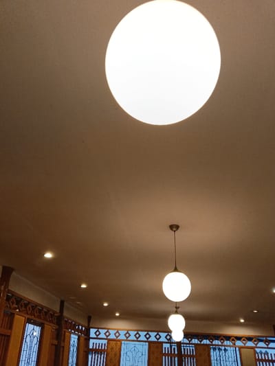 The image presents an upward-looking interior view of a building, focusing on its ceiling and upper architectural details. A dominant feature is a large, circular, bright white light fixture positioned prominently in the upper center of the frame, appearing to be a flush mount or large recessed light on the light brown ceiling. Below this, two spherical pendant lights hang at different heights, also emitting bright white light. The upper pendant is larger and more clearly visible than the lower one, which is partially obscured.

The ceiling also features several smaller, circular recessed spotlights scattered across its surface, contributing to the overall illumination of the space. In the lower part of the image, decorative wooden elements are visible, including intricate panels with geometric, diamond-shaped patterns and vertical wooden beams. Below these panels are windows or openings with dark grilles or patterned panes.

No people are present in the image, and no text is visible. The bright artificial lighting suggests an indoor setting, likely in the evening or at night, or an interior space designed to be brightly lit regardless of the time of day. The architectural style, particularly the decorative wooden panels, hints at a traditional or culturally-inspired design, aligning with a location such as Ciamis, Indonesia, possibly a restaurant, guesthouse, or a similar communal establishment.