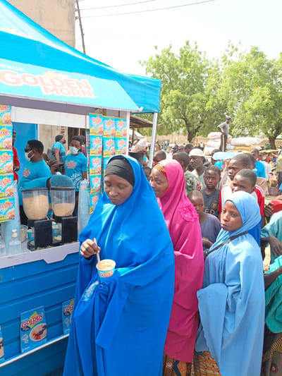 The image shows a crowd of people in Kawaji, Nigeria, gathered around a Nestle Golden Morn promotional stand. The stand is blue and white with Golden Morn branding and product displays. Three women in traditional Muslim clothing (blue, pink, and light blue hijabs) are in the foreground. The woman in blue is eating from a Golden Morn cup with a wooden spoon. Behind them, a diverse crowd of men, women, and children stand, many watching the activity at the stand. Some of the stand workers are wearing blue Golden Morn branded shirts and face masks. In the background, there are trees, buildings, and what appears to be a market or storage area with stacked sacks. The scene takes place during the day with bright sunlight.