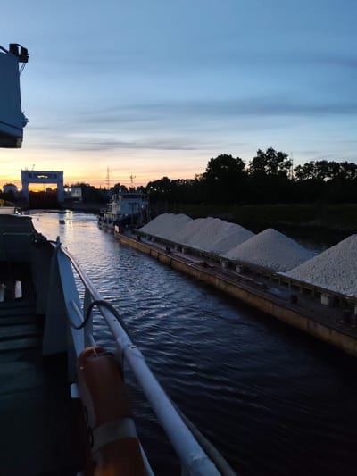 This image depicts a waterway, likely the Sheksna River, in Sheksna, Russia, at dusk. A large barge laden with white aggregate is being towed by a tugboat. The tugboat, with the name "МАЯК" (Mayak) visible on its side, is positioned behind the barge, moving forward. The camera is positioned on the deck of another vessel, looking forward and slightly to the right. In the background, a large lock structure is visible, illuminated by the fading light of sunset. Trees line the riverbanks, silhouetted against the sky. The sky displays a gradient of colors, from orange and yellow at the horizon to deep blue overhead, with thin clouds. The water is calm, with ripples reflecting the ambient light. A life preserver is attached to a railing in the foreground of the camera vessel. There are no people clearly visible in the image. The scene conveys a sense of industrial activity and transportation along the waterway.