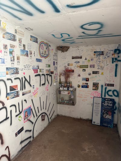 A small, white-walled corner space is filled with graffiti, stickers, and memorial elements. The walls are covered in Hebrew writing, drawings (including a sun), and spray paint. A glass case containing photos and small items sits on one wall, along with bottles. Flyers and brochures lean against the corner. A bird's nest is visible high on the wall. The space appears to be a makeshift memorial or tribute site, adorned with personal messages and images.