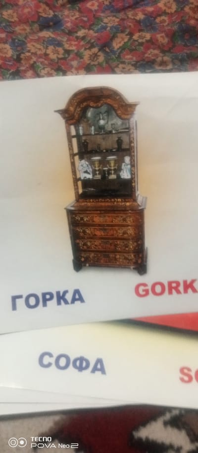 The image shows a miniature display cabinet filled with decorative items. The cabinet is dark brown with intricate gold detailing and sits on a white surface. Below the cabinet, there are words printed in blue and red. The blue text on the left reads "ГОРКА" and on the right, the red text reads "GORK". Below this, further to the left, blue text reads "СОФА". The bottom left corner shows the logo "TECNO POVA Neo 2". The background above the white surface is a floral pattern in shades of red, blue, and yellow.
The text suggests that this image may be related to a furniture store or catalog, possibly located in a city named "Tahmach, Uzbekistan" as indicated by the context. The items in the cabinet appear to be ornamental vases and figures. The overall scene is static and does not depict any action or interaction.